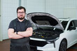 © pressmaster - Portrait of Caucasian middle aged man with beard standing with arms crossed in front of car with open hood in auto repair shop, looking directly at camera