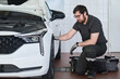 © pressmaster - Caucasian young adult man kneeling beside car inspecting front wheel in auto repair shop, wearing work gloves and glasses, focusing on maintenance task, vehicle hood open