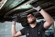 © pressmaster - Caucasian young adult man wearing glasses inspecting underside of vehicle with flashlight in auto repair shop, holding tool while examining car components during maintenance work