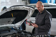 © pressmaster - Middle aged Caucasian man standing in auto repair shop using digital tablet while inspecting car engine, focusing on diagnostics and maintenance tasks in professional environment