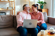 © DragonImages - Middle aged Caucasian man and middle aged Caucasian woman sitting on sofa embracing and laughing while using laptop together, teapot and cups on table in foreground