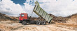© Anoo - At the construction site, a dump truck can be seen dumping earth onto the site, creating earthen embankments, while the sky is overcast. Heavy equipment is working on the site