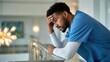 © SerPak - A male nurse in blue scrubs appears stressed while leaning on a railing, gazing out a window in a modern healthcare setting. Healthcare worker emotions.