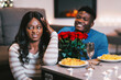 © Prostock-studio - Two people sit at a table with plates of pasta and roses. One person smiles while the other looks unsure. The setting is warm with soft lighting and decorations.