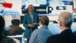 © DC Studio - African American female speaker delivers powerful media presentation at tech conference, diverse audience in modern conference hall. Young adult discusses advertising and public relations.