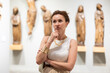 © JackF - Attractive woman in mid-thirties, with short brown hair, wearing white tank top and gold accessories, closely examining antique statues in art gallery, deeply engaged in understanding ancient origins