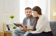 © Studio Romantic - Couple using tablet with discussion at home. Smiling wife pointing at screen with finger, man with beard looking at woman to discuss online news, search results, people sitting on living room sofa