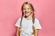 © SHOTPRIME STUDIO - Smiling young girl with blonde hair in braids wearing a white t-shirt stands against a pink background. She shows missing teeth and looks happy and playful. Studio portrait.