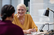 © Seventyfour - Middle aged Caucasian woman wearing glasses engaging in conversation with client in modern office setting, both sitting at desk with digital tablet visible