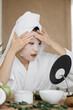 © sofiko14 - A young woman with a towel on her head applies a sheet mask to her face while wearing a white bathrobe and looking in a mirror