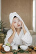© sofiko14 - A woman with a towel on her head and wearing a bathrobe applies a facial sheet mask while surrounded by fruits and skincare products