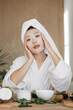 © sofiko14 - A young woman with a towel on her head and a white robe applies a sheet mask to her face while sitting at a table with skincare products and fruits