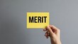 © Creative Parul  - Hand holding a bright yellow sticky note with the bold word 'MERIT', symbolizing achievement, recognition, and value on a grey background.