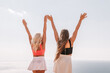 © svetograph - Two women are standing on a beach, one wearing a pink top and the other wearing a black top. They are both holding their hands up in the air, as if they are celebrating something. The sky is clear.