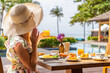 © TravelMedia - Woman in sun hat enjoying breakfast with orange juice at tropical resort poolside, surrounded by palm trees and summer ambiance, epitomizing relaxation and leisure travel.
