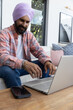 © wavebreak3 - Indian man working on laptop in living room on sofa, leaning forward with smartphone nearby