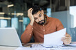 © Liubomir - Young male employee feeling tired and frustrated while looking at financial papers at his office desk, dealing with a deadline, stress, and professional burnout