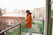 © H_Ko - A young woman stands on a modern balcony, wearing an orange coat and smiling gently. She is surrounded by vibrant city buildings, adding a lively atmosphere to the serene moment.