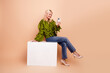 © deagreez - Stylish mature blonde woman model sits on white cube in green blouse and jeans using smartphone in a beige studio