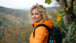 © Artsaba Family - A smiling woman with blonde curly hair enjoys a hike, wearing a bright orange puffer jacket and carrying a backpack