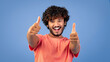 © Prostock-studio - A young man smiles broadly while giving two thumbs up. He wears a pink shirt and poses against a blue background. His happy expression shows excitement and positivity in the moment.