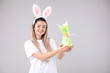 © New Africa - Easter celebration. Smiling woman in headband with bunny ears holding wrapped gift on grey background