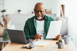 © Prostock-studio - A man sits at a desk in his home office. He looks at a document with a serious face while a laptop is open in front of him. Sunlight fills the room and plants are nearby.