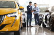 © Prostock-studio - Handsome middle eastern guy standing among brand new cars, having conversation with cheerful sales assistant, looking at catalogue in dealer hands, checking auto features, full length