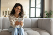 © Prostock-studio - Millennial Caucasian woman using smartphone while sitting on sofa, checking messages, having online video call at home, free space. Pretty young female posting in social media on mobile phone indoors