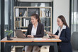 © Thitisak - Two businesswomen in professional suits working together, one holding a laptop and the other taking notes on a tablet, showcasing teamwork and technology in a modern corporate environment.