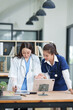 © Thitisak - female doctor in a white lab coat and a medical assistant in blue scrubs are sitting at a wooden desk, collaborating and reviewing medical documents or X-rays. Both are wearing stethoscopes, reflectin