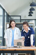 © Thitisak - female doctor in a white lab coat and a medical assistant in blue scrubs are sitting at a wooden desk, collaborating and reviewing medical documents or X-rays. Both are wearing stethoscopes, reflectin