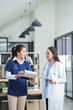 © Thitisak - female doctor in a white lab coat and a medical assistant in blue scrubs are sitting at a wooden desk, collaborating and reviewing medical documents or X-rays. Both are wearing stethoscopes, reflectin