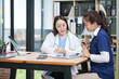 © Thitisak - female doctor in a white lab coat and a medical assistant in blue scrubs are sitting at a wooden desk, collaborating and reviewing medical documents or X-rays. Both are wearing stethoscopes, reflectin