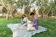 © WMSTUDIO - Young couple enjoys a relaxed afternoon in a serene park, playing ukulele and reading, surrounded by lush greenery and sunlight filtering through trees.
