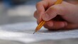 © Anastasia - Close-up of a person's hand holding a yellow pencil and writing on a piece of paper. the hand is positioned in a way that the pencil is pointing towards the right side of the image.