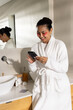 © Wavebreak Media - Mid-30s Asian woman standing at sink in white robe holding smartphone, wearing pink patches