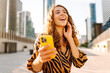 © maxbelchenko - A portrait of a smiling woman standing on a sunny city street with a phone. A beautiful woman blogs, enjoying a stroll overlooking the metropolis. Concepts for blogging and tourism.