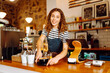 © maxbelchenko - A female barista stands behind the counter, serving freshly brewed coffee. A happy woman in an apron prepares aromatic coffee in a cozy coffee shop. Small business concept, drinks.