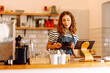 © maxbelchenko - A female barista stands behind the counter, serving freshly brewed coffee. A happy woman in an apron prepares aromatic coffee in a cozy coffee shop. Small business concept, drinks.