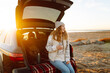 © maxbelchenko - A woman traveler holds a warm drink from a thermos in her hand while sitting in the trunk of a car at dawn. A young woman enjoys the seascape on a road trip. Concept of adventure, enjoyment.