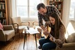 © Sergio - A man is showing a girl how to play guitar in a warm living room. They sit on a couch while paper and cups are on a table. Sunlight fills the space