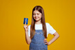 © Davidovici - Smiling kid holds a blue credit card with curiosity and excitement in front of a bright yellow backdrop. Ideal for financial education content and advertising related to children.