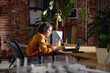© Seventyfour - Young adult woman sitting at desk working on laptop in modern office space, concentrating on screen, surrounded by plants and office supplies, exposed brick wall background