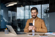 © Liubomir - Smiling man concentrating on his smartphone while sitting at a modern office desk with a laptop, reflecting modern work-life balance and efficient digital communication