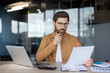 © Liubomir - Thoughtful businessman in glasses analyzing financial documents and reports, holding a pen to his chin while working at a desk with a laptop in a modern office, focusing on strategy and planning