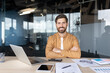 © Liubomir - Young male entrepreneur sitting at office desk with laptop and documents, smiling broadly at camera, portraying success, startup, and professional achievement