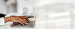 © Prostock-studio - Two hands are seen typing on a laptop at a bright workspace. The setting highlights focused work and productivity. Soft light fills the background creating a work environment.