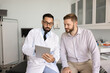 © fizkes - Health report review. Confident male doctor physician general practitioner in white coat hold digital tablet explain young man patient medical data lab test results on consultation in examination room