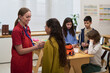 © pressmaster - Caucasian young adult woman using stethoscope examining Asian preteen girl, while diverse group of children sitting at table engaging with medical equipment in classroom setting